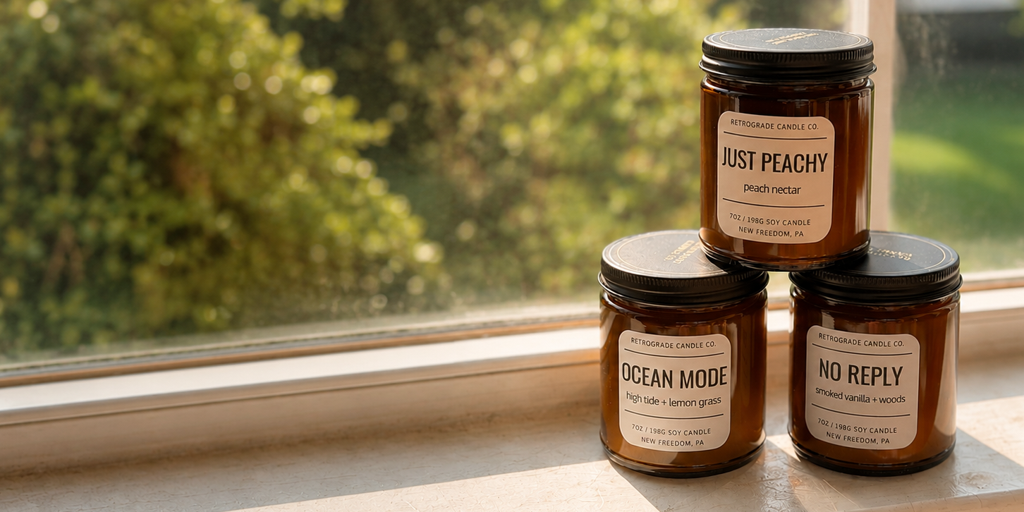 Three amber glass candle jars with black lids and white labels are stacked on a windowsill. The labels read 'JUST PEACHY', 'OCEAN MODE', and 'NO REPLY'.