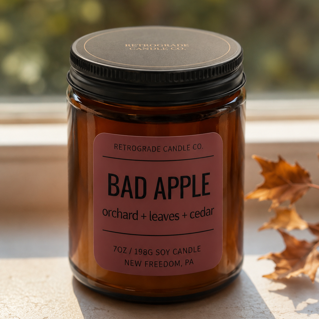 An amber glass jar candle with a black lid and a dusty pink label reading 'BAD APPLE orchid + leaves + cedar' by Retrograde Candle Co., sitting on a stone windowsill with warm natural light and autumn leaves nearby.