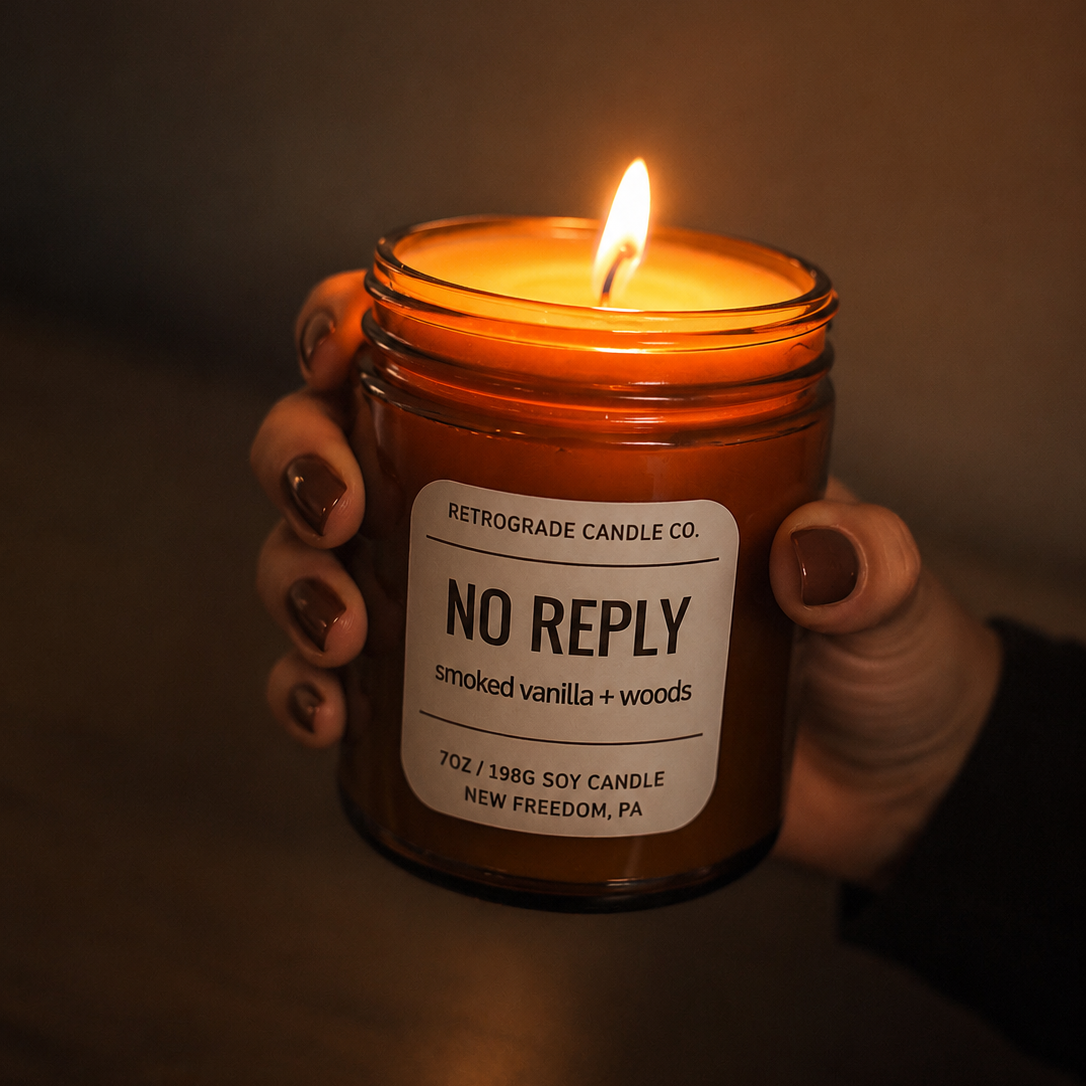 A hand with dark nail polish holds a lit amber glass jar candle labeled 'NO REPLY' by Retrograde Candle Co. against a dark, moody background.