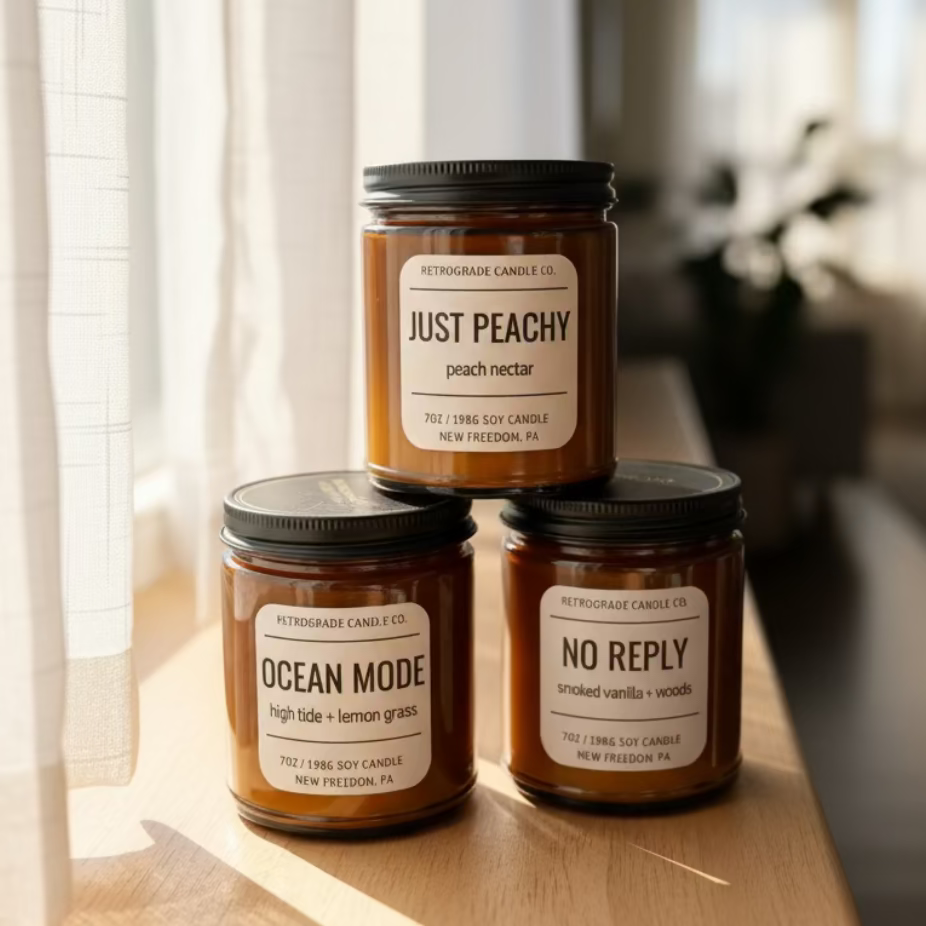 Three amber glass jar candles with black lids are arranged on a windowsill. The labels read Just Peachy, Ocean Mode, and No Reply, with Retrograde Candle Co.