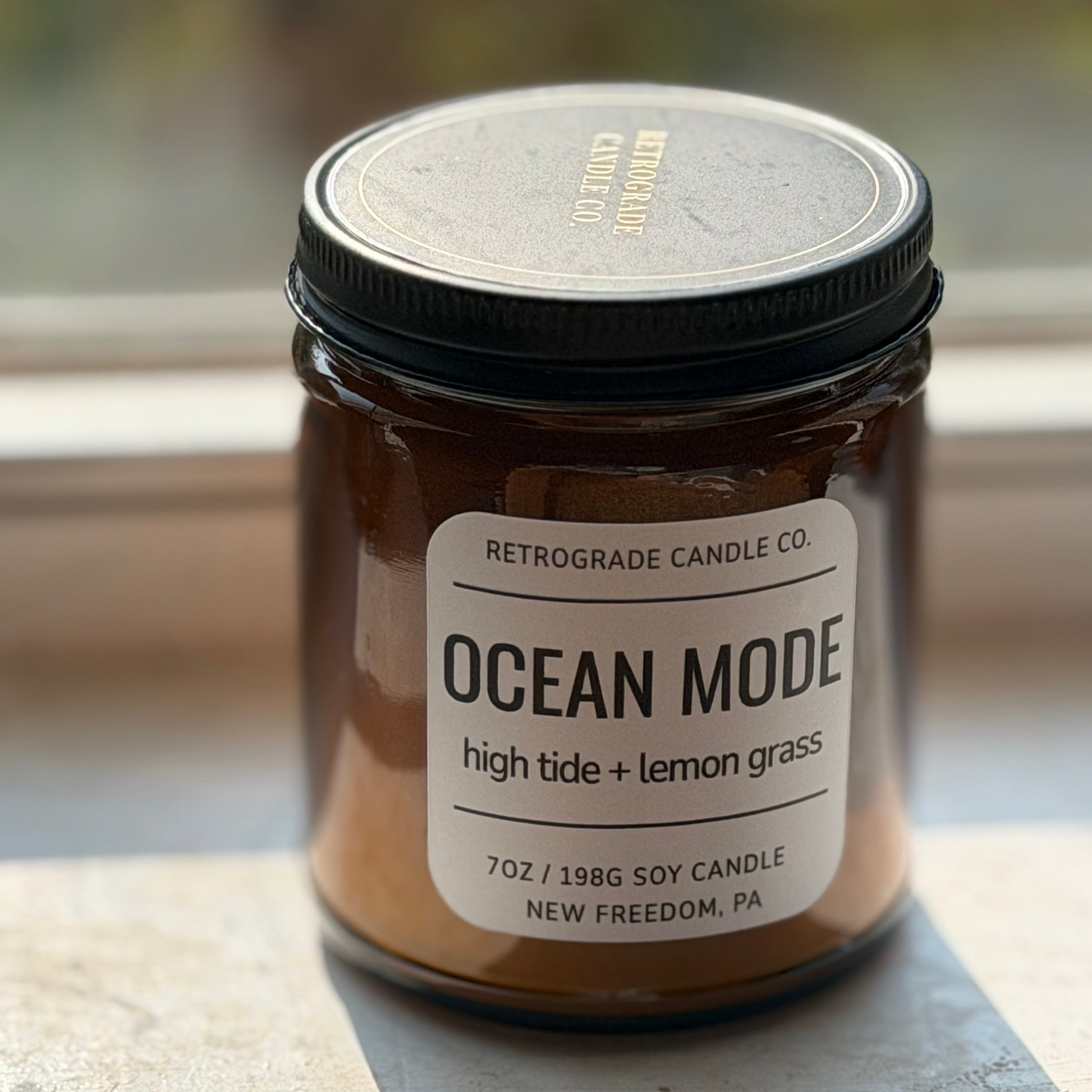 A 7oz soy candle in an amber glass jar with a black lid and a white label reading 'OCEAN MODE' by Retrograde Candle Co., sitting on a windowsill with natural light.