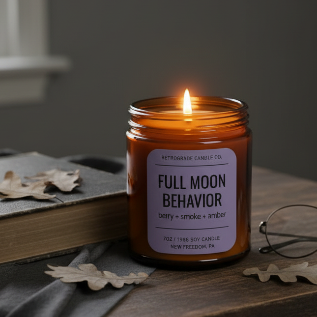 A lit amber glass jar candle on a dark wooden table, with a white label reading 'FULL MOON BEHAVIOR' and 'Retrograde Candle Co.' in warm ambient lighting.