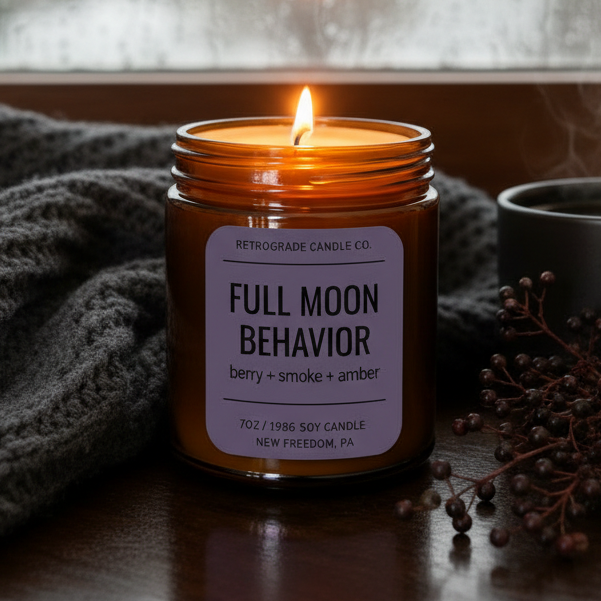 A lit amber glass jar candle on a dark wooden table, with a white label reading 'FULL MOON BEHAVIOR' and 'Retrograde Candle Co.' in warm ambient lighting.