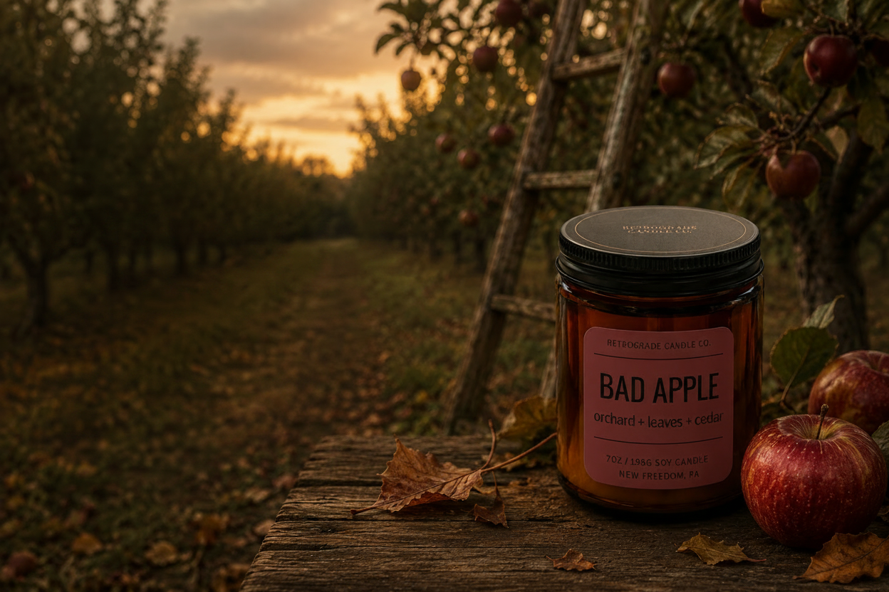 An amber glass jar candle labeled 'BAD APPLE' from the RCC Fall Collection sits on a rustic wooden surface outdoors in an apple orchard at sunset, with apples and leaves around it. Text on the left reads 'BAD APPLE' and 'RCC FALL COLLECTION'.